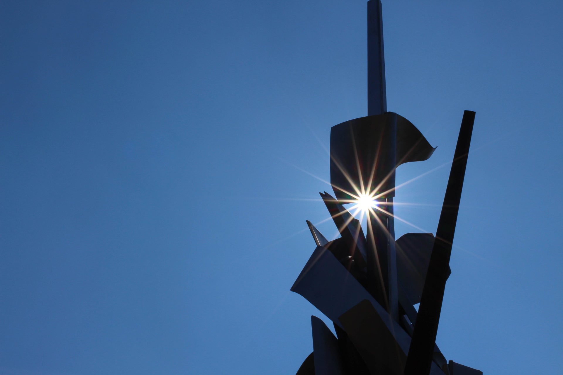 A wrought metal sculpture against a peerless blue sky. The sun shiens through a gap in the metal, casting a 14-point starburst.