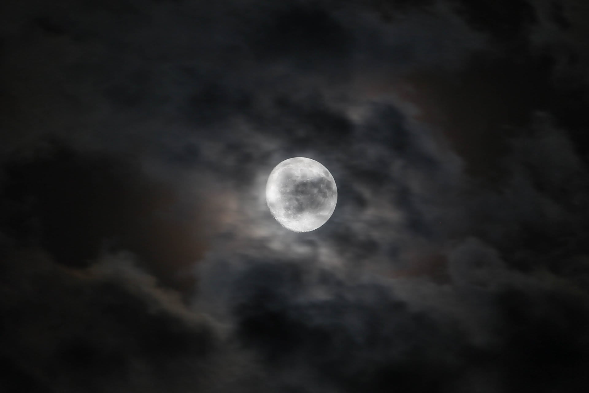 A waxing gibbous moon sits at the center of a hazy night sky, partially obscured by black clouds. The light of the moon throws the haze into a range of blurring browns, blacks, silvers, and greys.