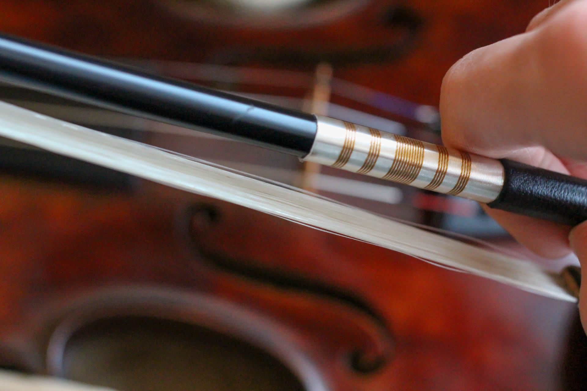A hand holding a carbon fiber bow, overtop the softly out-of-focus background of a deep reddish-brown violin.