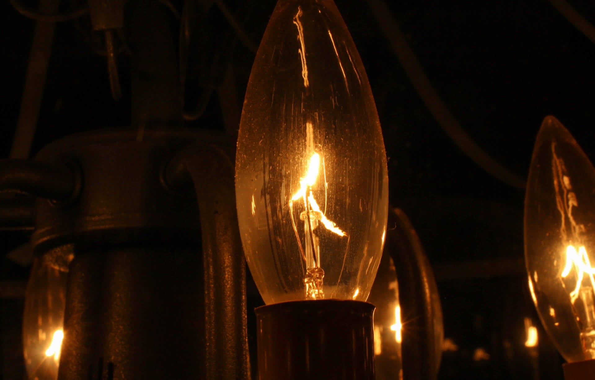 Yellow light burns across an electrical filament, encased inside an amber bulb. Matching bulbs and the curves of dark metal give the suggestion of a chandelier.