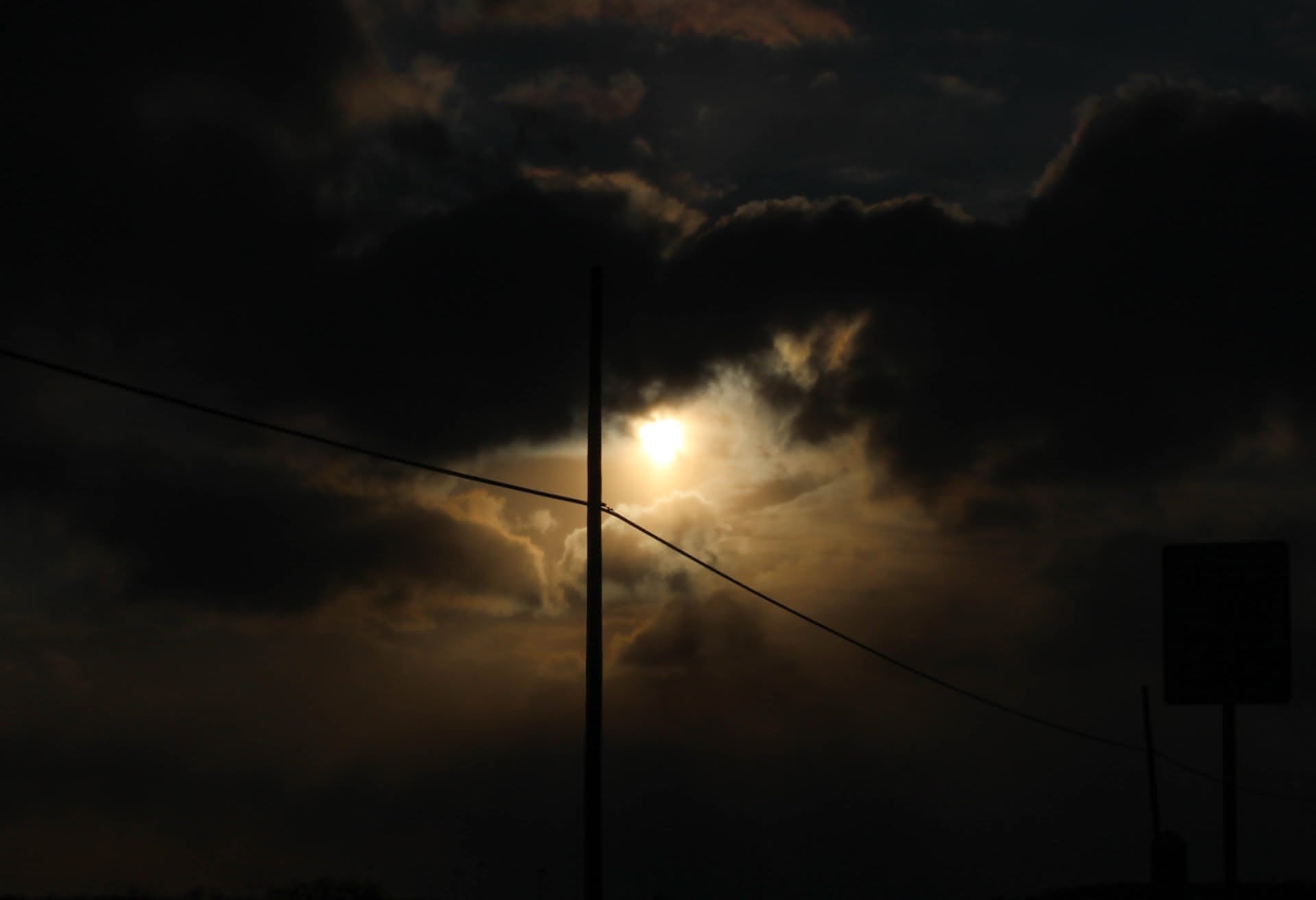 The silhouette of a power line crosses the scene, above which the sun peers out from amidst black and gold clouds like the eye of a dragon.