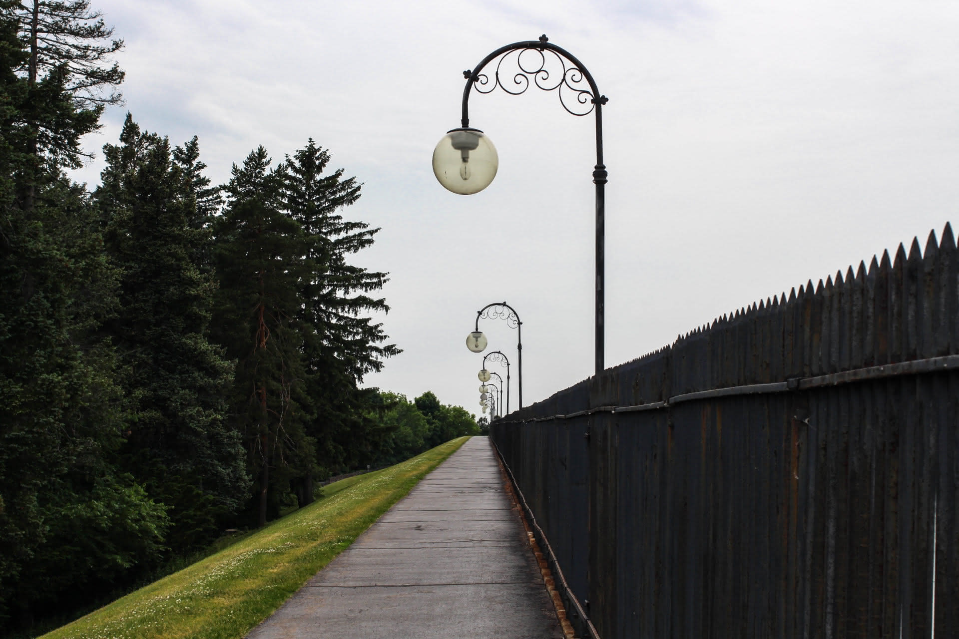 A straight cement path in 1-point perspective. A grassy hill leads down to dark green trees on the lefthand side, while the righthand side is enclosed by a black metal fence. Streetlamp bulbs arch over the path from the fence and vanish into the distance.