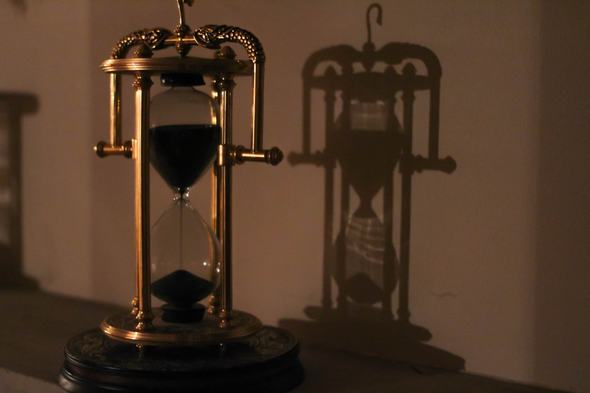 An ornate golden hourglass with black falling sand sits in soft focus, while its shadow casts a flattened impression upon the wall