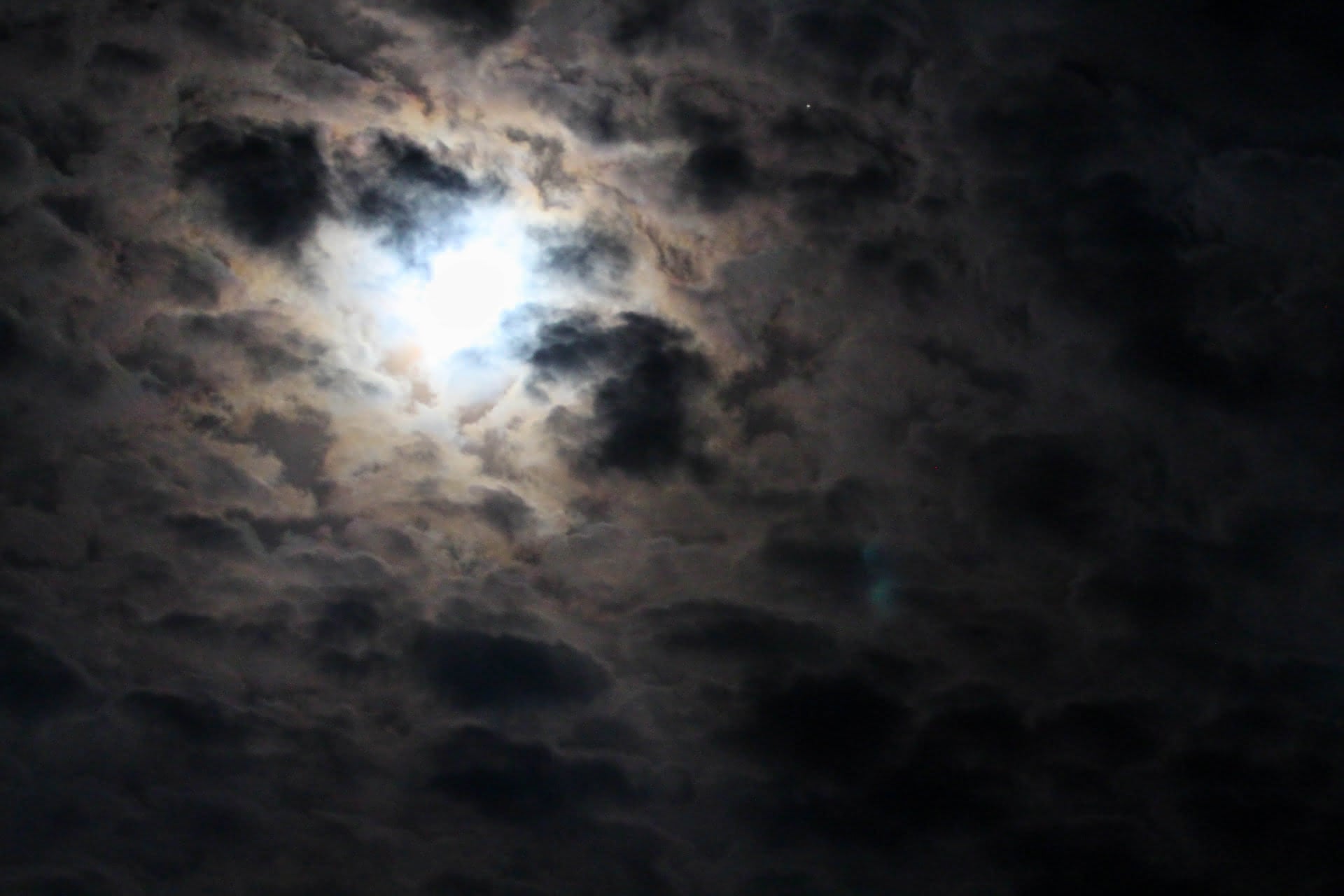 A brilliantly-illuminated night sky, where an ice white patch of moonlight solidifies the nearby clouds into almost resembling a cliff face of sharp black and tan rocks.