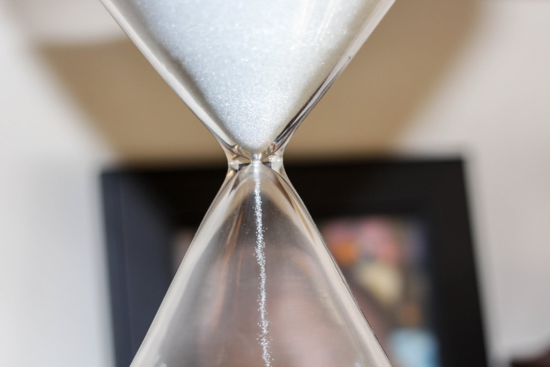 Tiny glowing grains of white sand fall from a close-cropped view of a plastic hourglass, with an out-of-focus black picture frame sitting at a dynamic angle in the background.