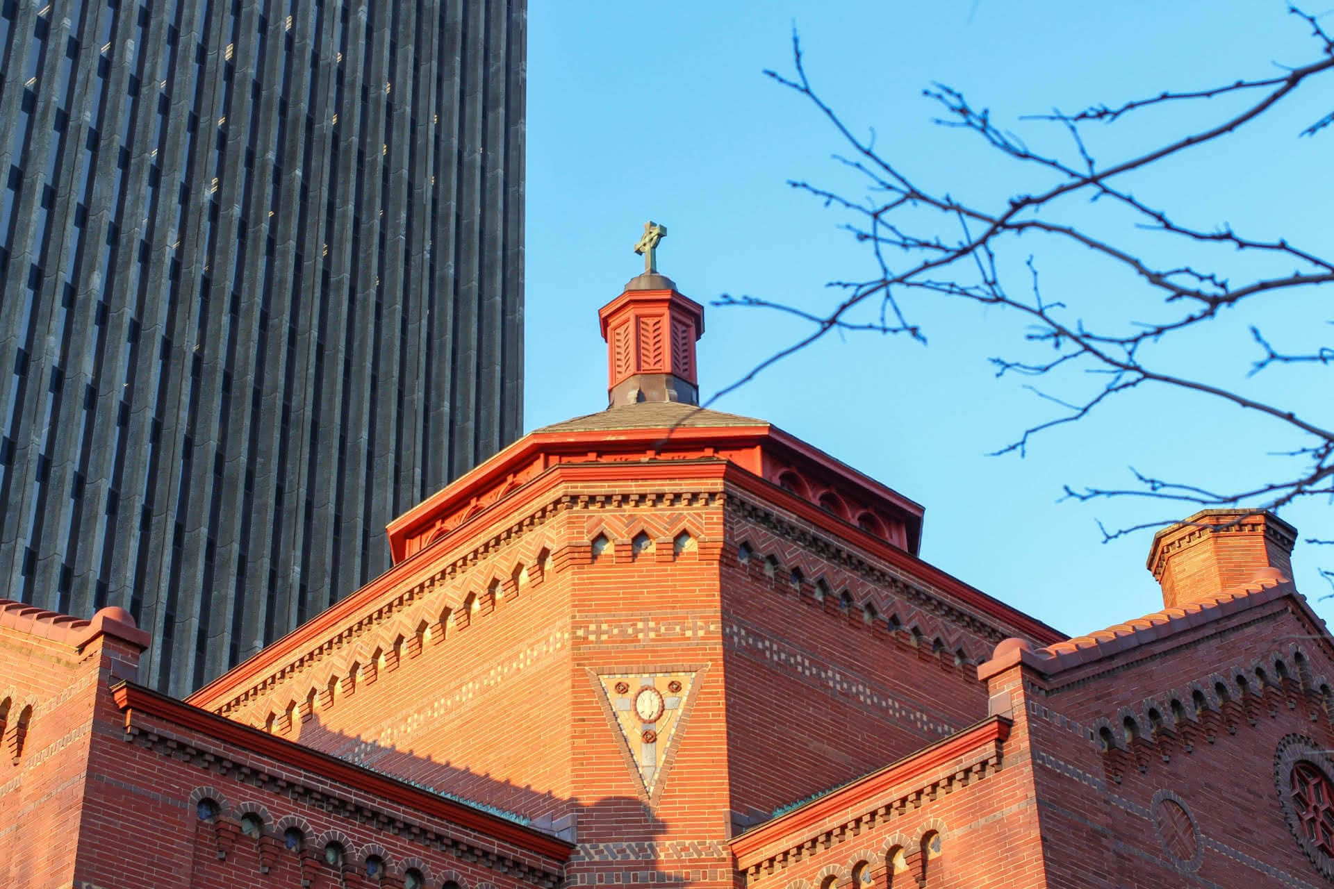 A brick church in the middle of a three-point composition, with a gray scyscraper to the left, and the branches of a tree to the right.